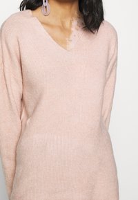 Maglione a maglia di un morbido rosa con scollo a V, texture a costine e vestibilità leggermente oversize. Presenta maniche lunghe e dettagli di cucitura in vista.
