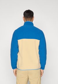 Μπλε και μπεζ φλις μπουφάν με ψηλό γιακά, σχέδιο με χρωματικά μπλοκ και ελαστική ζώνη στο τελείωμα. Μαλακή υφή και άνετη, καθημερινή εφαρμογή.