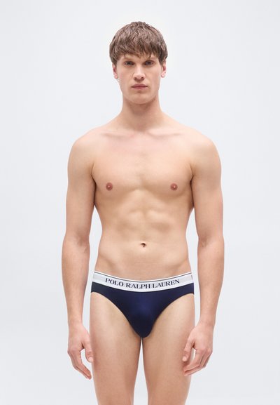 Σκούρα μπλε ανδρικά εσώρουχα από απαλό ύφασμα με λευκή ζωγραφισμένη λωρίδα στη μέση. Διαθέτουν εφαρμοστή σχεδίαση και ελάχιστη κάλυψη, κατάλληλα για άνδρες.