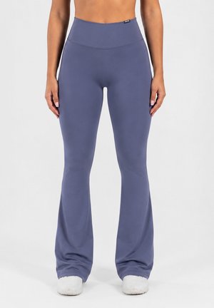 Mujer con pantalones de yoga azules de talle alto y acampanados y zapatillas blancas, de pie con los brazos relajados a los lados.