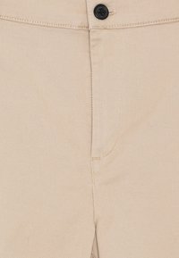 Gros plan sur un pantalon beige montrant la fermeture par bouton, la ceinture et le contour de la poche cousue sur un tissu sergé.