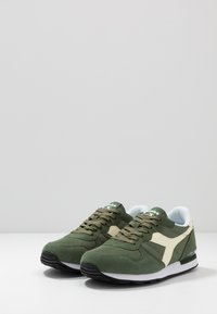 Green athletic shoes featuring a mix of textured suede and mesh, with cream accents and black rubber soles. Light blue interior lining.