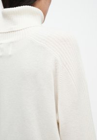 Λευκό πουλόβερ με ψηλό γιακά και ριγωτή υφή στους ώμους, με απαλή και ελαφριά αίσθηση.