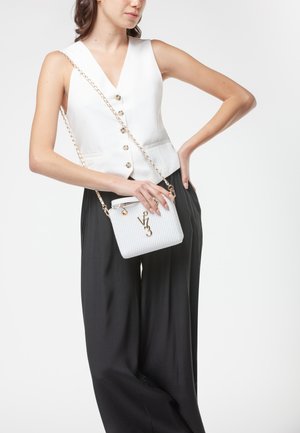 Donna che indossa un gilet bianco senza maniche con bottoni e pantaloni neri a gamba larga, che tiene una piccola borsa a tracolla bianca intrecciata con tracolla a catena dorata.