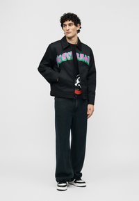 Νεαρός με σγουρά μαλλιά που φοράει μαύρο μπουφάν με πολύχρωμο κείμενο "ICECREAM", μαύρο γραφικό μπλουζάκι, φαρδιά σκούρα τζιν και μαύρα-άσπρα αθλητικά παπούτσια.