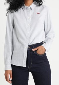Camicia a righe blu e bianca con logo rosso sul petto, abbinata a jeans scuri a vita alta. Collo standard, maniche lunghe.