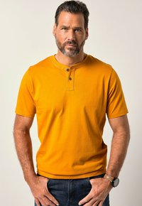 Maglietta giallo senape a maniche corte con scollatura henley, due bottoni neri e una texture liscia. Abbinata a jeans scuri.