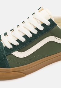 Låg sneaker med överdel av mörkgrön mocka och tyg, vit läderdetalj i vågform, vita snören och sula av gummirubber.