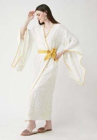 White kimono-style robe with a textured pattern, yellow trim, and sash. Loose sleeves, reaching ankle length. Brown slide sandals.