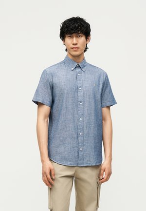 Jonge man draagt een blauw overhemd met korte mouwen en beige cargobroek, staand voor een effen lichte achtergrond.