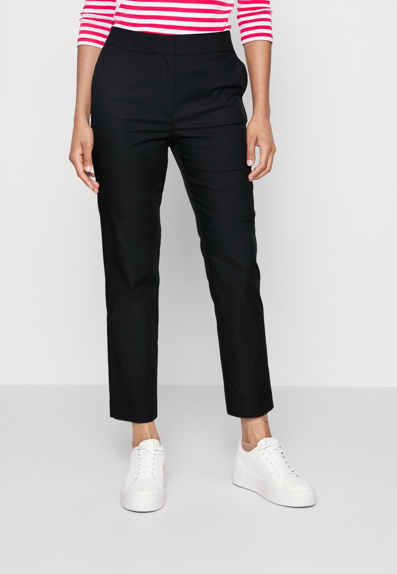 Black, tapered pants with a smooth texture and elastic waistband, paired with white sneakers. Minimalist design, no visible patterns.