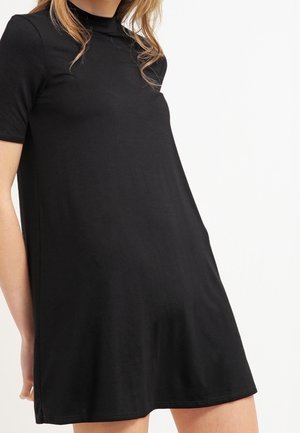 Femme portant une robe noire à manches courtes avec un col montant, debout devant un fond blanc uni.