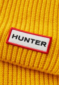 Žuti pleteni šešir s crvenom ivicom i oznakom "HUNTER" u crnom tekstu. Tekstura je rebrasta s vidljivim pletenim uzorcima.