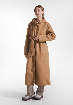 Frau steht und trägt einen langen braunen Kapuzenmantel, der in der Taille gebunden ist, hellgraue Hose und graue Sportschuhe vor einem einfarbigen hellen Hintergrund.