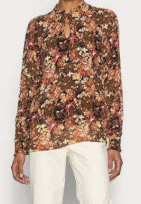 Blouse à manches longues dans un motif floral marron, avec un décolleté en forme de clé, en tissu léger et coupe décontractée, associée à un pantalon de couleur crème.
