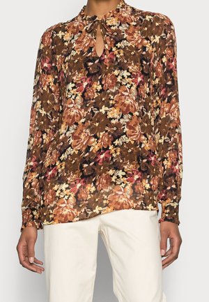 Langärmlige Bluse im braunen Blumenmuster, mit einem Schlitz-Ausschnitt, leichtem Stoff und lockerer Passform, kombiniert mit cremefarbenen Hosen.