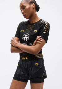 Črni komplet športnih oblačil UFC, ki vsebuje teksturirano majico s kratkimi rokavi z zlatimi poudarki in logotipom, ter ujemajoče se hlače s potiskom in oprijetim pasom.