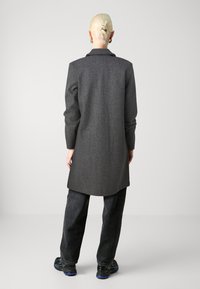 Šedý kabát z vlněné směsi s volnějším střihem, dlouhými rukávy a klopovým límcem, doplněný tmavými džínami a černými sportovními botami.