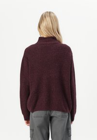 Sweater en maille bordeaux avec un col montant, coupe ample et texture côtelée. Associé à un pantalon cargo gris, présentant une silhouette décontractée.