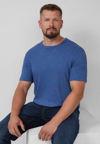 Korte mouwen T-shirt met ronde hals, van blauw gemêleerd stof met een subtiele textuur, gecombineerd met donkere jeans. Model zit tegen een neutrale achtergrond.