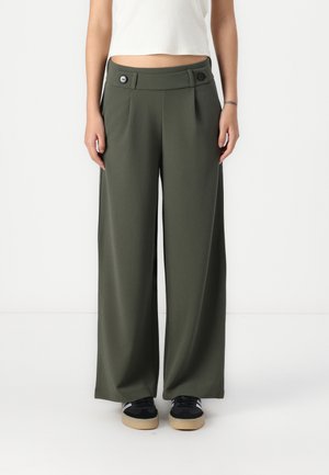 Personne portant un pantalon taille haute, à jambes larges, couleur vert olive, avec deux boutons à la taille, associé à des baskets noires et un top court blanc.