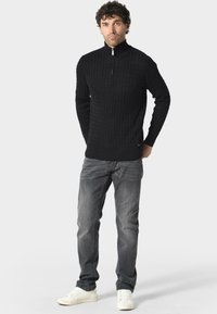 Czarny dziany sweter z wysokim kołnierzem i zamkiem błyskawicznym na ćwierć długości, z teksturowanym wzorem. W połączeniu z szarymi dżinsami i białymi trampkami.