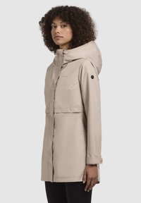 Beige waterdichte parka met capuchon, ritssluiting aan de voorkant en een patch pocket. Heeft een relaxte fit en knoopaccenten op de mouwen.