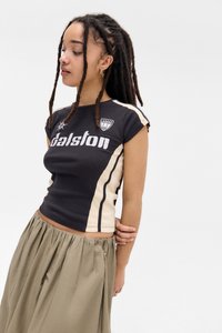 Jauna sieviete ar gariem dreads, tērpusies melnā un bēšā Dalston t-kreklā un olīvzaļās svārkās, skatās uz leju pa kreisi.