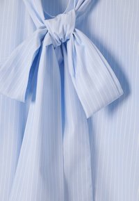 Light blue blouse with vertical white stripes. Features a soft, textured fabric and a tied neck detail with flowing ends.
