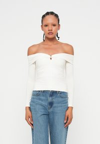 Off-the-shoulder white top with long sleeves, ruched design, and a front keyhole detail featuring a small metallic accent. Paired with blue jeans.