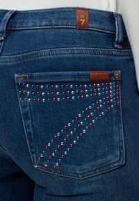 Poche arrière d’un jean en denim bleu décorée de clous rouges, blancs et bleus formant un motif stylisé du chiffre sept et une petite étiquette en tissu.