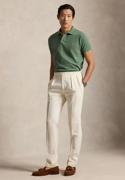 Vyras vilki žalią trumpomis rankovėmis polo marškinėlį, kreminius klostuotus kelnes, rudus kutuotus loaferius ir laikrodį ant riešo, stovintis priešais vienspalvį smėlio spalvos foną.