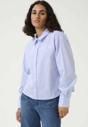 Chemise à rayures bleues avec un col et une poche poitrine, associée à un jean bleu ajusté. Tissu texturé avec des boutons blancs en accent.