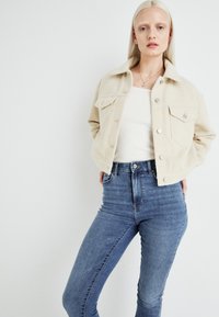 Beige steppelt dzseki gombos elülső résszel és mellzsebekkel, magas derekú kék farmernadrággal párosítva, sima textúrával, testhezálló kivitelben.