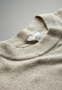 Béžový vlnený sveter s rebrovaným okrúhlym výstrihom. Má mäkkú textúru a štítok s názvom značky a veľkosťou.