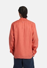 Camicia di lino corallo con colletto classico, maniche lunghe e polsini con bottone. Presenta un gancio sulla schiena per appenderla. Texture liscia e design leggero.