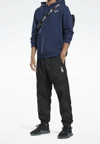 Personne portant un sweat à capuche Reebok navy, un pantalon de survêtement Reebok noir, des baskets noires et un sac bandoulière noir avec des sangles blanches portant un logo.