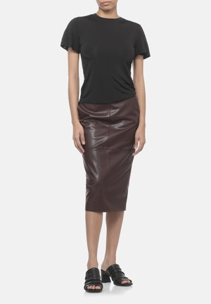 Donna con top nero a maniche corte, gonna a matita in pelle marrone scuro e sandali neri con tacco e punta aperta, in piedi davanti a uno sfondo semplice.