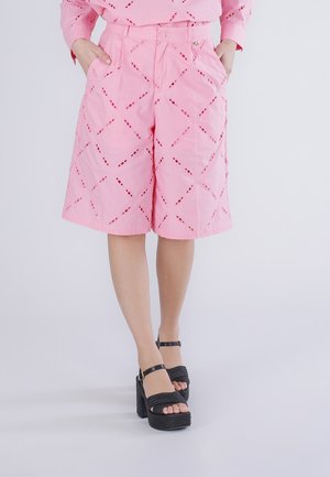 Kvinde iført knælange shorts med pink mønster, med hænderne i lommerne og sorte højhælede platformsandaler på en ensfarvet baggrund.