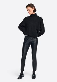 Schwarzer, texturierter Rollkragenpullover kombiniert mit eleganten schwarzen Lederhosen und schwarzen Loafers, vor einem hellen Hintergrund.