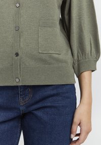Cardigan verde lavorato a maglia con scollatura rotonda, bottoni frontali, una tasca singola e polsini a costine, abbinato a jeans in denim blu scuro.