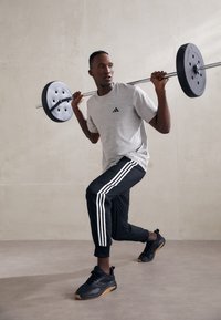 Pelēks īsām piedurknēm krekls ar melnu Adidas logotipu, melnas sporta bikses ar baltām svītrām un melnas sporta apavi. Cēliena svarus ar stieni, uz kura ir svari.