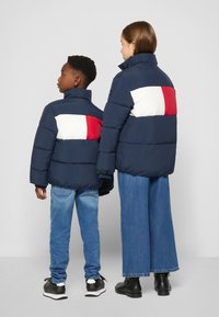 Marina puffjackor med vita och röda horisontella ränder på ryggen, kombinerat med ljusblå jeans och svarta skor. Inga synliga logotyper.