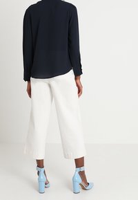 Nainen pukeutuneena mustaan pitkähanteiseen puseroon, valkoisiin leveälahkeisiin keskipitkiin housuihin ja vaaleansinisiin korkokenkiin, seisoo selkä kameralle.