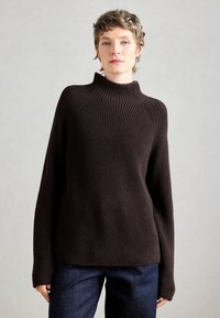 Maglione a maglia marrone scuro con colletto alto e texture a coste. Vestibilità ampia con maniche lunghe e taglio dritto all'orlo. Indossato con jeans scuri.