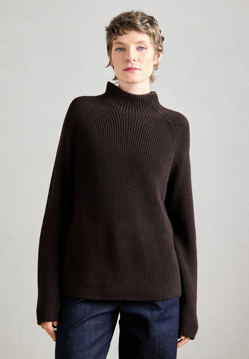 Maglione a maglia marrone scuro con colletto alto e texture a coste. Vestibilità ampia con maniche lunghe e taglio dritto all'orlo. Indossato con jeans scuri.