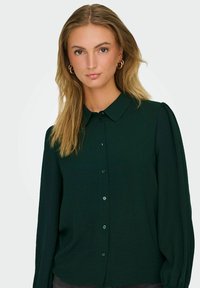 Dunkelgrünes Hemd mit Knopfleiste aus strukturiertem Stoff, klassischem Kragen und langen Ärmeln, die über feine Rüschen an den Schultern verfügen.