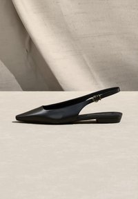 Μαύρο δερμάτινο παπούτσι χαμηλού τακουνιού με λουράκι στο πίσω μέρος, μυτερή μύτη και μικρή πόρπη σε μπεζ επιφάνεια και φόντο.