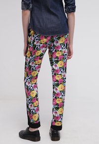 Bloementrousers met een zwarte achtergrond, versierd met levendige roze, gele en blauwe bloemenpatronen. Afgemaakt met zwarte enkelmanchetten en gedragen met zwarte schoenen.