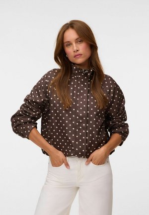 Femme aux longs cheveux bruns portant une veste marron à pois blancs et un pantalon blanc, debout les mains dans les poches devant un fond blanc.